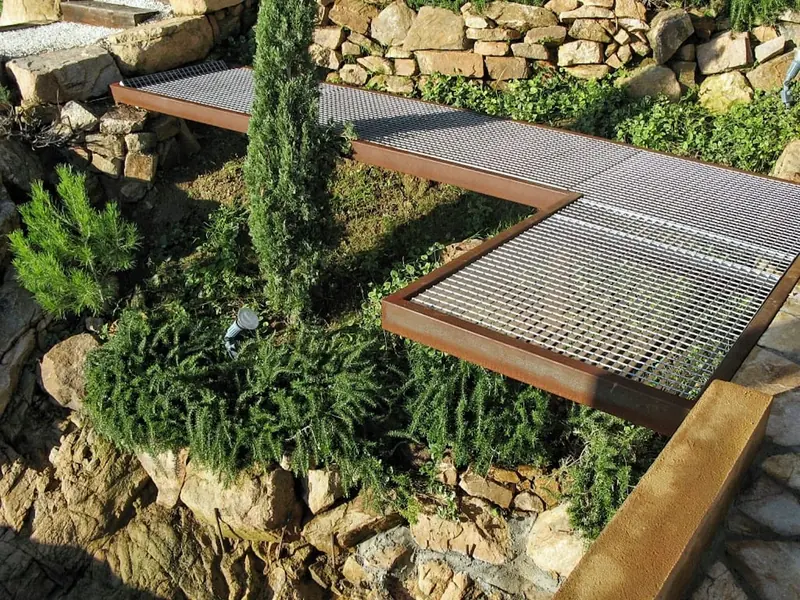 Passarel·la de reixa metàl·lica i acer corten sobre un talús amb plantes aromàtiques