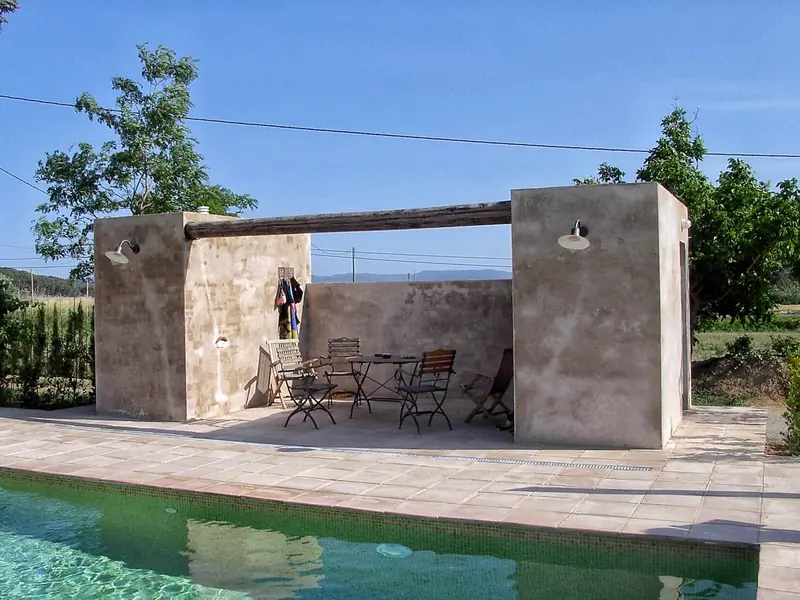 Pavelló d’estuc amb pèrgola davant d’una piscina, amb taula i cadires de jardí