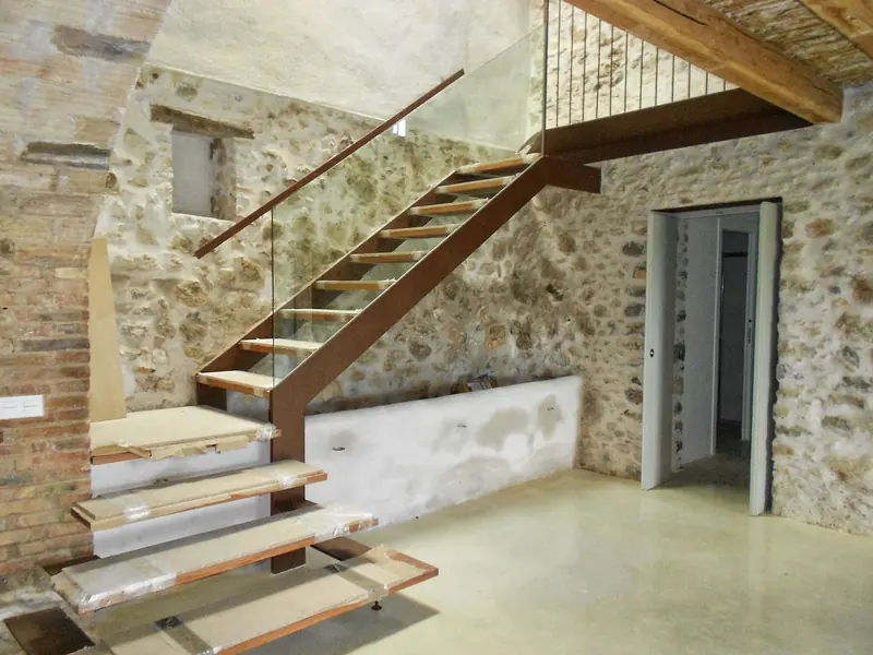 Escala de fusta i vidre en un interior de pedra vista, en fase d’acabats