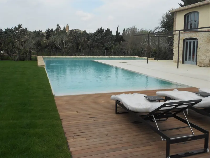 Solàrium de fusta amb gandules al costat d’una piscina, davant d’una casa de pedra